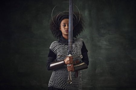 Photo for Brave young African woman, medieval warrior in chainmail armor with halo-like rings above head holding sword close to face on dark vintage background. Concept of history, emotions, comparison of eras - Royalty Free Image
