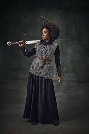 Photo for Young African-American woman medieval warrior in armor holding sword on shoulder, standing against dark vintage background. Concept of history, beauty and fashion, comparison of eras - Royalty Free Image