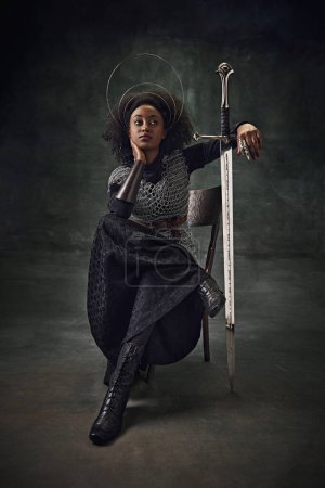 Téléchargez les photos : Belle jeune fille africaine, guerrière médiévale en armure de maille avec des anneaux halo au-dessus de la tête assise sur une chaise, appuyée sur l'épée. Concept de comparaison des époques, histoire, art créatif, remake - en image libre de droit