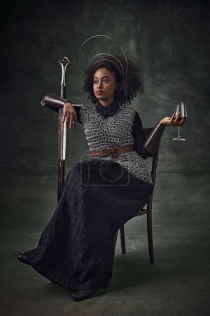Foto de Jovencita africana, guerrera medieval con armadura de cota de malla sentada en silla con espada y copa de vino sobre fondo verde vintage. Concepto de comparación de épocas, historia, arte creativo, remake - Imagen libre de derechos