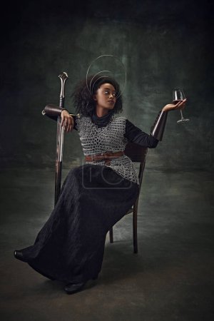 Téléchargez les photos : Jeune fille africaine élégante, guerrière médiévale en armure de maille assise sur une chaise avec épée et verre à vin sur fond vert vintage. Concept de comparaison des époques, histoire, art créatif, remake - en image libre de droit