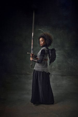 Téléchargez les photos : Jeune femme africaine, guerrière médiévale en armure de mailles avec des anneaux de halo au-dessus de la tête et plume d'ange sur le dos tenant l'épée. Ange noir. Concept de comparaison des époques, histoire, remake - en image libre de droit