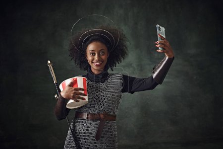 Photo for Smiling young African woman, medieval warrior in chainmail armor holding sword and taking selfie with mobile phone against vintage green background. Concept of comparison of eras, history, remake - Royalty Free Image