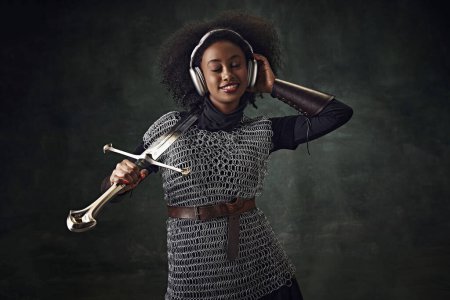 Téléchargez les photos : Jeune femme africaine souriante, guerrière médiévale en armure de mailles avec épée écoutant de la musique dans des écouteurs sur fond vert vintage. Concept de comparaison des époques, histoire, modernité, remake - en image libre de droit