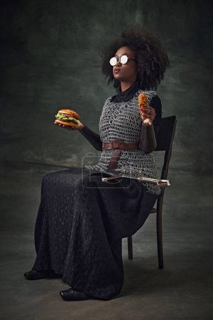 Téléchargez les photos : Femme africaine, guerrière médiévale en cotte de maille, lunettes de soleil, hamburger et nuggets de poulet sur fond sombre vintage. De la malbouffe. Concept de comparaison des époques, histoire, art créatif, remake - en image libre de droit