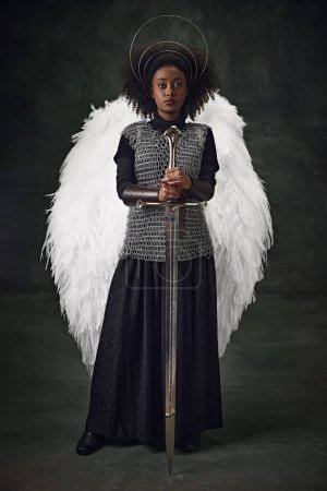 Téléchargez les photos : Jeune femme africaine aux ailes d'ange, halo au-dessus de la tête et maille tenant l'épée sur fond sombre vintage. Concept de comparaison des époques, histoire, art créatif, remake - en image libre de droit