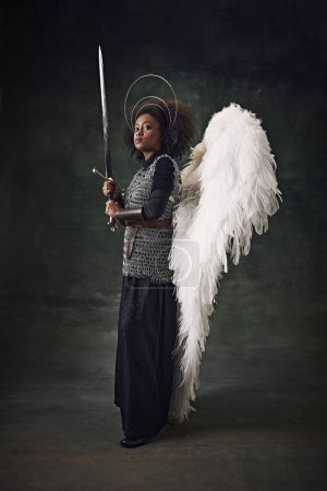 Photo for Young African woman, medieval warrior with angel wings, halo above head and chainmail holding sword against vintage dark background. Concept of comparison of eras, history, creative art, remake - Royalty Free Image