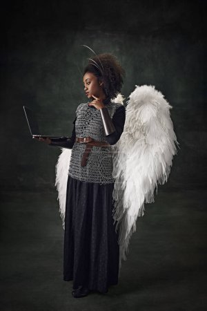 Foto de Joven mujer africana, guerrera medieval con alas de ángel sosteniendo portátil contra fondo oscuro vintage. Tecnologías modernas. Concepto de comparación de épocas, historia, arte creativo, remake - Imagen libre de derechos