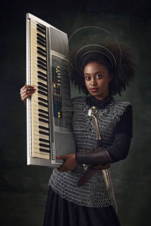 Téléchargez les photos : Jeune femme africaine, guerrière médiévale en chainmail et halo tenant clavier électronique sur fond vert vintage. Concept de comparaison des époques, histoire, art créatif, remake - en image libre de droit