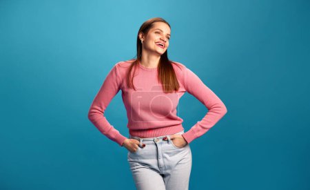 Photo for Portrait of beautiful young brunette girl in pink sweater and jeans smiling, laughing against blue studio background. Concept of youth, lifestyle, casual fashion, human emotions - Royalty Free Image