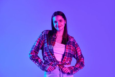 Photo for Smiling beautiful woman in checkered shirt posing against purple studio background in neon light. Positive vibe. Concept of youth, lifestyle, casual fashion, human emotions - Royalty Free Image
