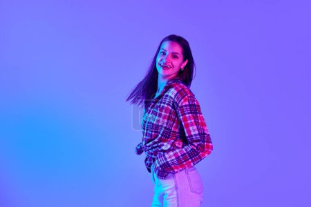 Foto de Mujer joven feliz y tierna con camisa a cuadros y jeans bailando y divirtiéndose contra el fondo del estudio púrpura con luz de neón. Concepto de juventud, estilo de vida, moda casual, emociones humanas - Imagen libre de derechos