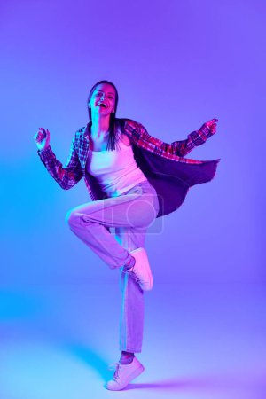 Photo for Happy and positive young woman in checkered shirt cheerfully dancing and having fun against purple studio background in neon light. Concept of youth, lifestyle, casual fashion, human emotions - Royalty Free Image