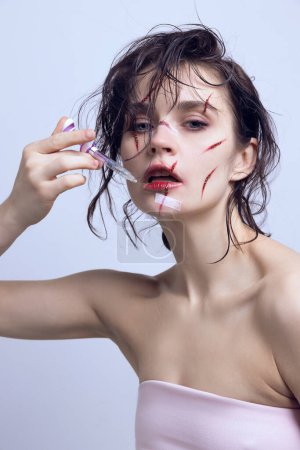Victims of beauty in modern ages. Beautiful young woman with syringe on lips and scratches on face. Concept of modern beauty standards, plastic surgery, health, cosmetology