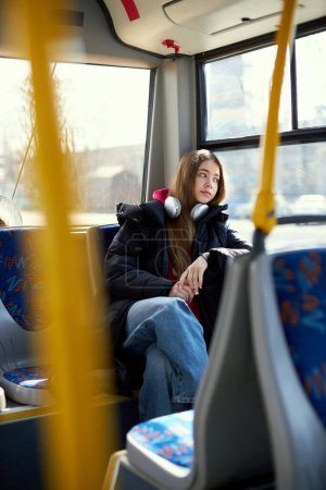 Téléchargez les photos : Jeune femme rêveuse dans un casque assis dans un tramway moderne. Transport sûr. Concept de transport public, mode de vie urbain - en image libre de droit