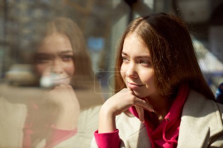 Téléchargez les photos : Jeune fille à capuche rose assise dans les transports en commun, tramway moderne, regardant par la fenêtre. Journée ensoleillée. Concept de transport public, mode de vie urbain - en image libre de droit