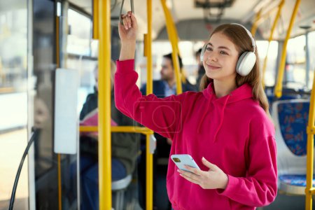 Téléchargez les photos : Jeune fille en sweat à capuche rose debout dans les transports en commun, tramway moderne, souriant, écoutant de la musique dans les écouteurs. Journée ensoleillée. Concept de transport public, mode de vie urbain - en image libre de droit
