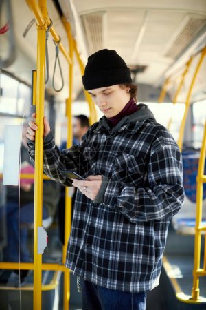 Téléchargez les photos : Jeune homme en chemise à carreaux debout dans le tramway moderne, bus et regardant sur le téléphone mobile. Mode de transport pratique et bon marché. Concept de transport public, mode de vie urbain - en image libre de droit