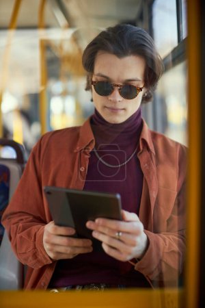 Téléchargez les photos : Jeune homme élégant dans des lunettes de soleil assis sur le tramway moderne et regardant sur tablette, lecture de livres, nouvelles. En utilisant les transports publics. Concept de transport public, mode de vie urbain - en image libre de droit