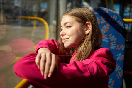 Foto de Jovencita con capucha rosa sentada en transporte público, tranvía moderno, autobús, mirando por la ventana y sonriendo. Viaje de un día soleado. Concepto de transporte público, estilo de vida urbano - Imagen libre de derechos