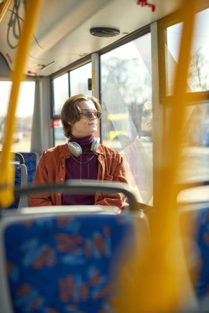 Photo for Young stylish man in sunglasses and headphones sitting on modern tram and looking in window. Using public transport. Sunny day. Concept of public transport, urban lifestyle - Royalty Free Image