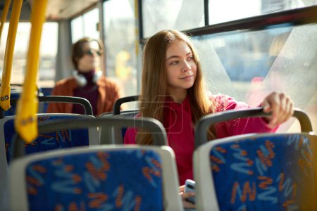 Téléchargez les photos : Focus sur la jeune fille en sweat à capuche rose assise sur le tramway avec passager flou sur le fond. Manière pratique pour le transport. Concept de transport public, mode de vie urbain - en image libre de droit