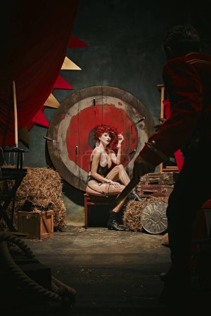 Photo for Young redhead woman with makeup and costume sitting and smoking near target over dark retro circus backstage background. Man throwing knives. Concept of circus, performance, show, retro and vintage - Royalty Free Image