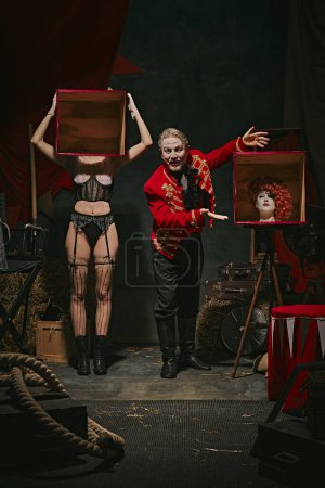 Photo for Tricks. Magician in red coat showing tricks with headless female assistant holding boxes over dark retro circus backstage background. Concept of circus, theater, performance, show, retro and vintage - Royalty Free Image
