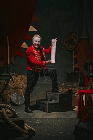 Magician with pale face makeup, in red coat making performance with cards over dark retro circus backstage background. Concept of circus, theater, performance, show, retro and vintage