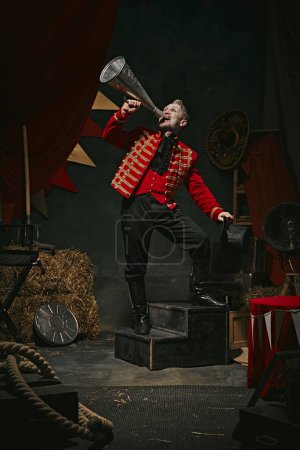 Photo for Announcement. Poster. Ringmaster, magician in red coat shouting into megaphone over dark retro circus backstage background. Concept of circus, theater, performance, show, retro and vintage - Royalty Free Image