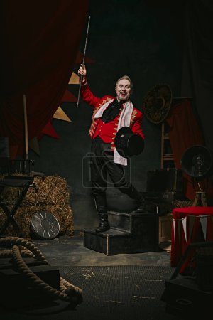Hombre con maquillaje de cara pálida, en traje de escenario rojo y sombrero haciendo rendimiento creativo sobre fondo retro oscuro circo entre bastidores. Concepto de circo, teatro, performance, espectáculo, retro y vintage