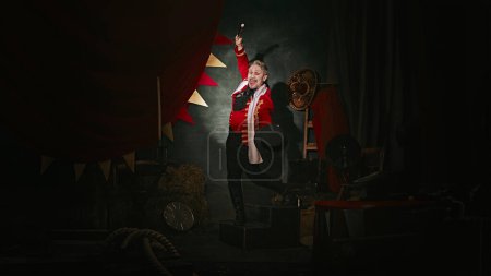Hombre emocional con maquillaje de cara pálida, en traje de escenario rojo y sombrero haciendo rendimiento sobre fondo oscuro circo retro tras bastidores. Concepto de circo, teatro, performance, espectáculo, retro y vintage