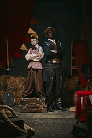 Photo for Retro-themed event. High African man standing with little servant boy over dark retro circus backstage background. Concept of circus, theater, performance, show, retro and vintage - Royalty Free Image