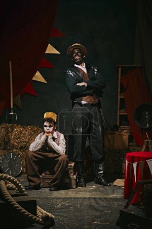 Photo for High African man with dirty face standing with little servant boy over dark retro circus backstage background. 20th-century vaudeville era. Concept of circus, theater, performance, retro and vintage - Royalty Free Image