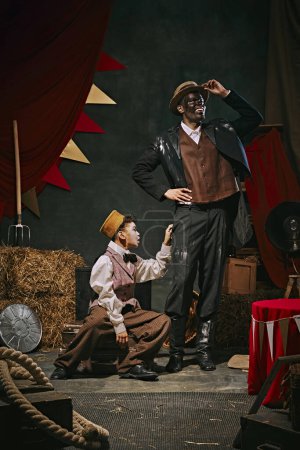 Photo for High smiling African man with dirty face standing with little servant boy over dark retro circus backstage background. Concept of circus, theater, performance, show, retro and vintage - Royalty Free Image