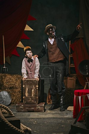 Photo for High African man with dirty face and clothes standing with little servant boy over dark retro circus backstage background. Concept of circus, theater, performance, show, retro and vintage - Royalty Free Image
