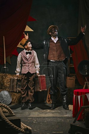 Photo for High African excited man with dirty face standing with little servant boy over dark retro circus backstage background. Concept of circus, theater, performance, show, retro and vintage - Royalty Free Image