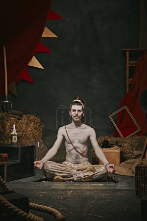 Photo for Pale man, yogi sitting in meditating pose over dark retro circus backstage background. Creative performance. Concept of circus, art, theater, performance, show, retro and vintage - Royalty Free Image