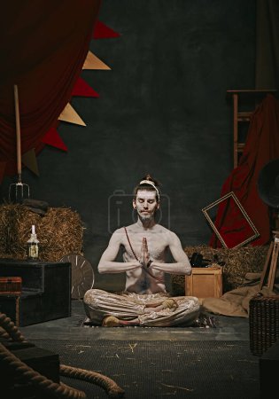 Photo for Pale man, yogi sitting in meditating pose over dark retro circus backstage background. Creative performance. Concept of circus, art, theater, performance, show, retro and vintage - Royalty Free Image