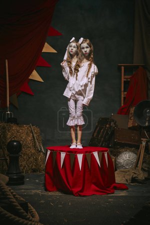 Siamesische Zwillingsmädchen in weißen Vintage-Kostümen mit Make-up stehen auf der Bühne vor dunklem Retro-Zirkus-Hintergrund. Gruselige Zaubershow. Konzept aus Zirkus, Theater, Performance, Retro und Vintage