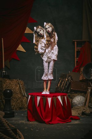 Siamese twin girls in white vintage costumes with makeup standing on stage over dark retro circus backstage background. Concept of circus, theater, performance, show, retro and vintage