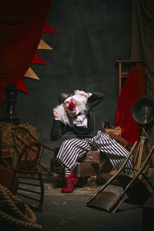 Clown in black hat, white face with red nose and striped pants shouting over dark retro circus backstage background. Concept of circus, theater, performance, show, retro and vintage