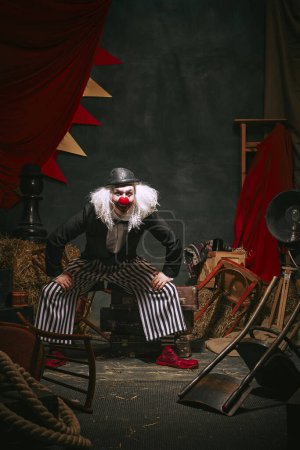 Clown mit schwarzem Hut, weißem Gesicht mit roter Nase und gestreifter Hose spielt vor dunklem Retro-Zirkus-Hintergrund. Komiker. Konzept aus Zirkus, Theater, Performance, Show, Retro und Vintage