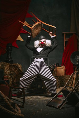 Téléchargez les photos : Clown au chapeau noir, visage blanc avec nez rouge et pantalon rayé rendant la performance avec chaise sur fond de coulisses de cirque rétro foncé. Concept de cirque, théâtre, performance, spectacle, rétro et vintage - en image libre de droit