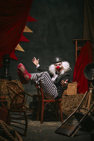 Foto de Payaso en sombrero negro, cara blanca con nariz roja y pantalones a rayas que actúan sobre fondo retro oscuro del circo entre bastidores. Juego emocional. Concepto de circo, teatro, performance, espectáculo, retro y vintage - Imagen libre de derechos