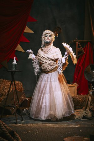 Foto de Drag queen en vestido blanco y maquillaje brillante sobre fondo de circo retro oscuro detrás del escenario. Hombre en apariencia femenina. Concepto de circo, teatro, performance, espectáculo, retro y vintage - Imagen libre de derechos