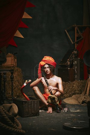 Photo for Young man, charmer in turban sitting with flute and cobra emerging from basket over dark retro circus backstage background. Concept of circus, theater, performance, show, retro and vintage - Royalty Free Image