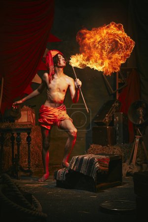 Photo for Fire show. Young man in turban breathes fire, displaying dramatic flame burst over dark retro circus backstage background. Concept of circus, theater, performance, show, retro and vintage - Royalty Free Image