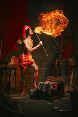 Photo for Fire show. Young man in turban breathes fire, displaying dramatic flame burst over dark retro circus backstage background. Concept of circus, theater, performance, show, retro and vintage - Royalty Free Image