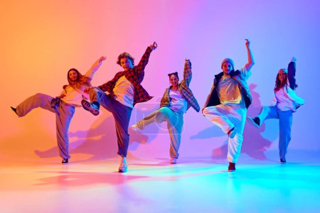 Téléchargez les photos : Performance dynamique de cinq danseurs talentueux en mouvement, dansant la danse moderne sur fond de studio dégradé sous la lumière du néon. Concept de style de danse moderne, passe-temps, mode de vie actif, culture des jeunes - en image libre de droit